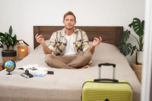 Portrait of young man meditating on a bed after packing suitcase for summer holiday, sitting with crossed legs in zen, nirvana pose, relaxing, hoping for something. Tourism and mindfulness concept