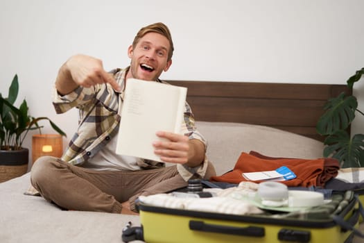 Portrait of young man pointing at his list of items, packing clothes on vacation, showing notebook, sitting near opened suitcase. Lifestyle and people concept