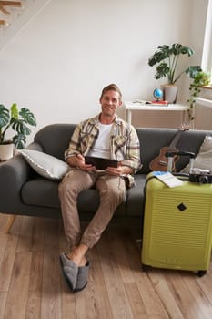 Portrait of handsome young man, tourist sitting with digital tablet near suitcase and two tickets, booking something online for his vacation, preparing for holiday.