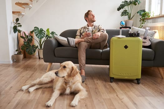 Portrait of happy, handsome young man with suitcase, sitting with his dog in living room, prepared to go on vacation, going on holiday. Tourism concept
