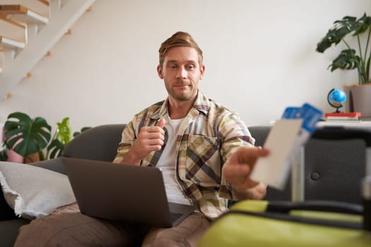 Portrait of happy young man booking holiday for two, has plane tickets, looks at laptop, sits in living room with computer. Travelling and tourism concept