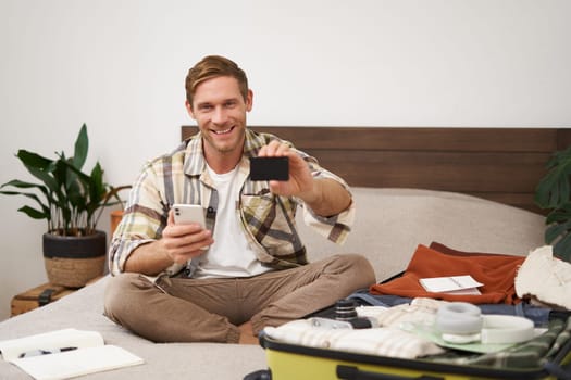 Portrait of happy man buying tickets with credit card cashback, sitting on bed and packing clothes for holiday, putting items in suitcase, holding mobile phone.