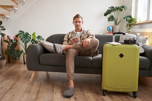 Portrait of man sits with mobile phone in living room, packed suitcase, chats on smartphone app, looks for holiday destinations, goes on vacation.