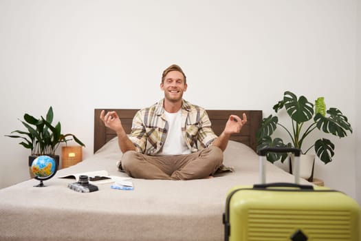 Portrait of young relaxed tourist, man sitting on bed with crossed legs and meditating, has packed luggage, globe and camera, going on holiday, calms down before going on a trip.