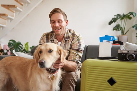 Happy young man with a dog, goes on holiday, packs suitcase and tickets, pets golden retriever. Pet-friendly hotels and tourism concept