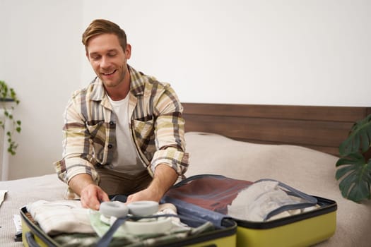 Portrait of young man, tourist going travelling abroad, putting clothes in suitcase, closing his luggage, sitting on bed, getting ready for holiday. Tourism concept