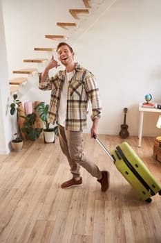 Vertical portrait of happy young man going on holiday, shows mobile phone call hand gesture, walking with suitcase, leaving rented apartment. Travelling and tourism concept