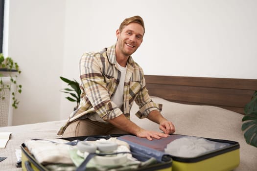 Portrait of young handsome bearded guy, tourist going on holiday, fastening his clothes in suitcase, packing for vacation, going on business trip, sitting in bedroom.