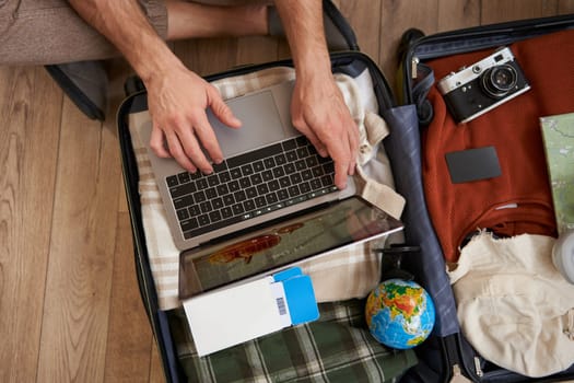 Close up portrait of male hands using laptop on top of opened suitcase, man packing for holiday, tourist with globe, booking vacation on travel agency website, picking a tour. Tourism concept