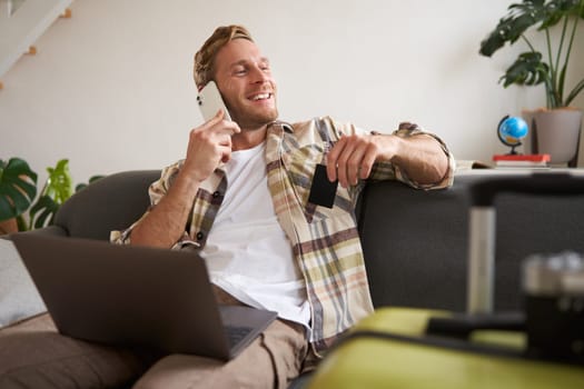Smiling guy talking on mobile phone, sitting with laptop, holding credit card, looking out of the window with happy, relaxed face expression.