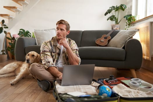 Portrait of young man, traveller going on holiday with a dog, packing his suitcase, sitting with laptop on his luggage, thinking, making online booking.
