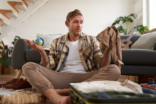 Portrait of confused man with suitcase, holding pants, shrugging shoulders and looking clueless at clothes luggage.