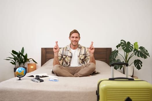 Portrait of young man meditating on a bed after packing suitcase for summer holiday, sitting with crossed legs in zen, nirvana pose, relaxing, hoping for something. Tourism and mindfulness concept