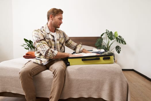 Portrait of young man packing his luggage for a trip, going on holiday, sitting on bed and putting clothes in suitcase, preparing for his flight.