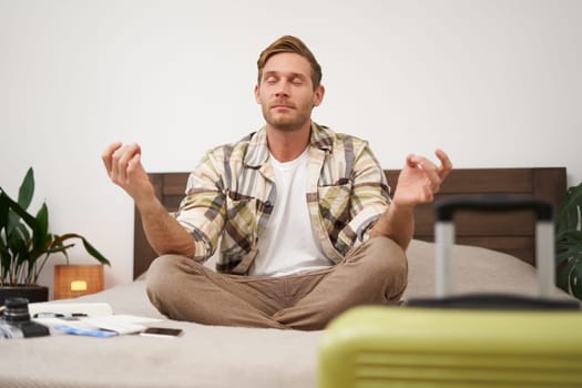 Close up portrait of meditating tourist, man sits with suitcase in bedroom, practice yoga, relaxing, finding nirvana. Tourism and travelling concept