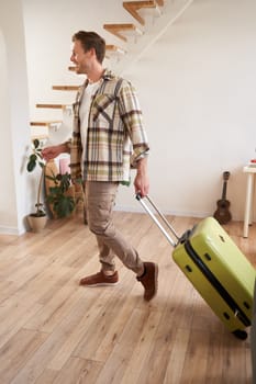 Full length photo of young man with suitcase, going on holiday trip, walking in flat, checking out from rented apartment. Copy space