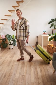 Vertical portrait of happy friendly man, guy waves hand at camera and says hello or bye, goes on vacation, leaves flat with suitcase. Concept of travelling, tourism and holiday.
