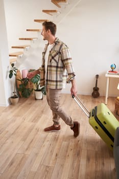 Vertical portrait of young handsome man walking indoors with suitcase, going on vacation, leaving his rented apartment with luggage.