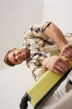 Image of funny man trying to close the suitcase filled with pile of clothes, packing luggage, putting things inside the bag. Tourism and traveller concept