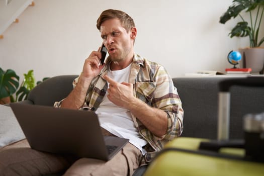 Portrait of angry man arguing over the phone, has laptop opened on his laptop, sits near suitcase, complaining at travel agency. Copy space