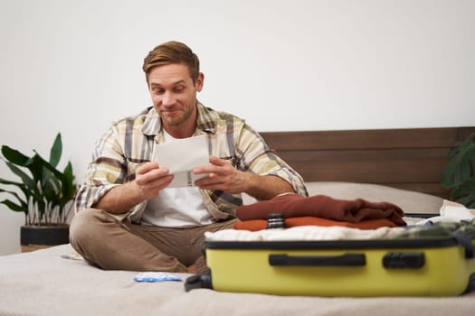 Portrait of excited young man, tourist going on vacation, has his tickets, sitting near opened suitcase, looking at passport with pleased smile. Lifestyle concept