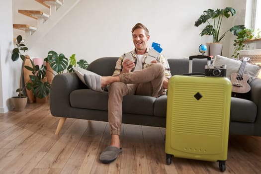 Portrait of man sits with mobile phone in living room, packed suitcase, chats on smartphone app, looks for holiday destinations, goes on vacation.