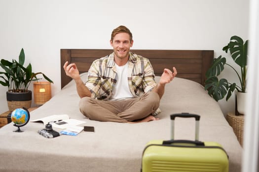 Portrait of young man meditating, packing suitcase, sitting on bed with globe, camera and plane tickets, relaxing before travelling abroad. Tourism concept