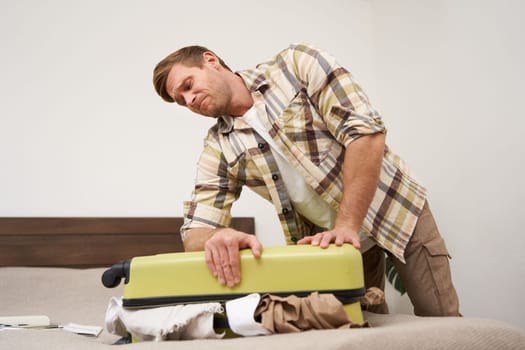 Side view of young man pushing on suitcase, trying to close it, has lots of clothes sticking out of luggage, going on long holiday, packing for vacation. Travelling and tourism concept