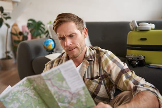 Portrait of happy young man, tourist with suitcase, sits in living room and looks at map, chooses holiday destination, picks route for travelling.