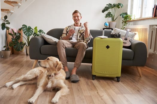 Happy man with his dog, sits in living room with suitcase, looks at smartphone app with excited face, pleased with booked trip. Tourism and travelling concept