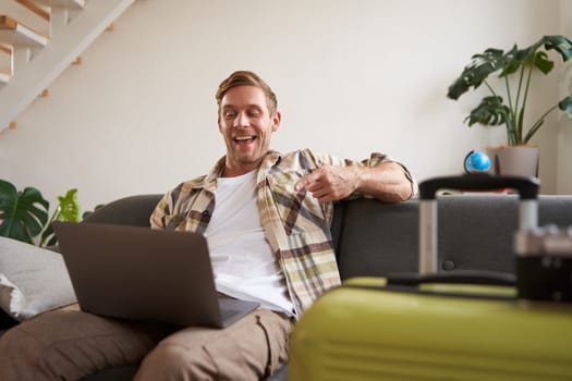 Happy handsome guy, sits with laptop, points finger at screen and laughs, has packed suitcase and camera prepared for holiday, goes on vacation. Tourism and travelling concept
