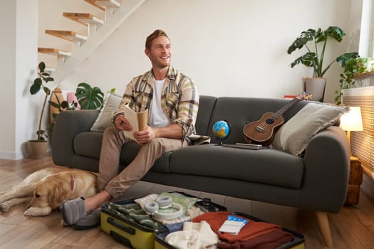 Portrait of man sitting with his dog, packing suitcase for a holiday, going on trip with a pet. Tourism concept