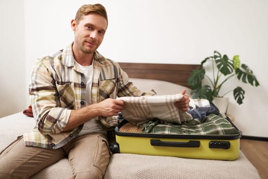 Portrait of man, tourist in hotel, packing or unpacking his suitcase, holding shirt, taking clothes out of luggage bag, smiling at camera.