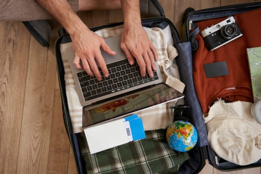 Close up of tourist, hands typing on laptop, opened suitcase with clothes for holiday, man packs luggage.