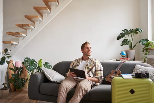 Image of young man, tourist goes on holiday, packed suitcase, sits with digital tablet on sofa, reads, looks at his map, books hotel for business trip. Copy space