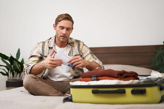 Portrait of handsome man, tourist, looking at his holiday tickets, booked vacation with travel agency, sitting on bed and packing clothes in suitcase.