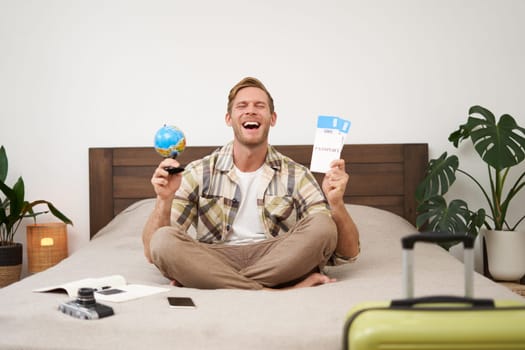 Portrait of cheerful, happy tourist, man sitting with plane tickets and a globe, packed suitcase for travelling around the world, going on vacation, excited about his holiday.