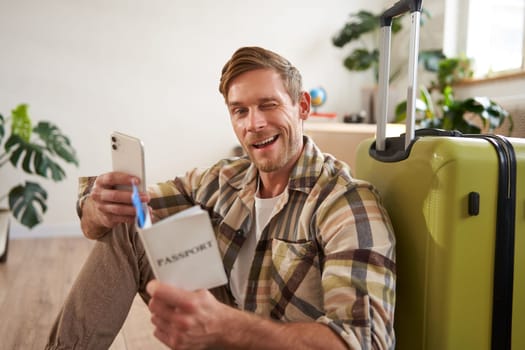 Charismatic young man, traveller going on holiday, trip abroad, sitting with suitcase, holding smartphone, tickets and passport, winking and smiling at camera.