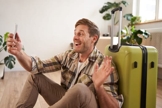 Cheerful smiling man with suitcase, video chats using smartphone, takes selfie, goes on holiday.