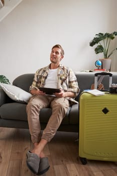 Image of young man, tourist goes on holiday, packed suitcase, sits with digital tablet on sofa, reads, looks at his map, books hotel for business trip. Copy space