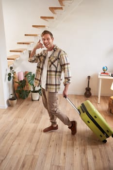 Full length shot of happy traveller, tourist goes on vacation, walks with suitcase, shows smartphone call gesture, leaves the apartment. Copy space