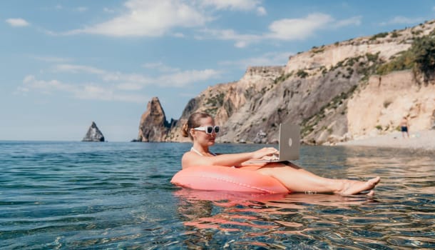 Woman freelancer works on laptop swimming in sea on pink inflatable ring. Pretty lady typing on computer while floating in the sea on inflatable donut at sunset. Freelance, remote work on vacation