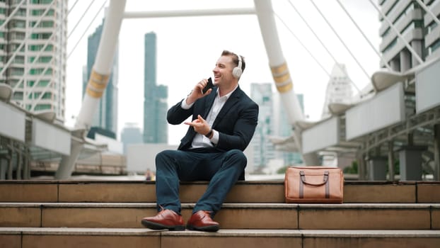 Professional business man listening headphone and using phone record voice while sitting at stair in urban city. Manager using headset listening relaxed song and moving along in lively mood. Urbane.