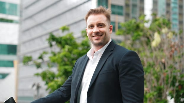 Skilled caucasian business man looking at camera while standing at rooftop. Professional investor smiling with confident while wearing formal suit at modern building in green city. Lifestyle. Urbane