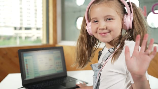 Smiling girl looking while waving hand at camera with laptop placed on table. Child wearing headphone smiling while laptop screen show system programing or coding program in STEM class. Erudition.