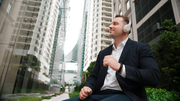 Low angle camera of project manager listening relaxing music while move to song and clap hands. Happy caucasian businessman enjoy listening music while sitting at bench at green urban city. Urbane.