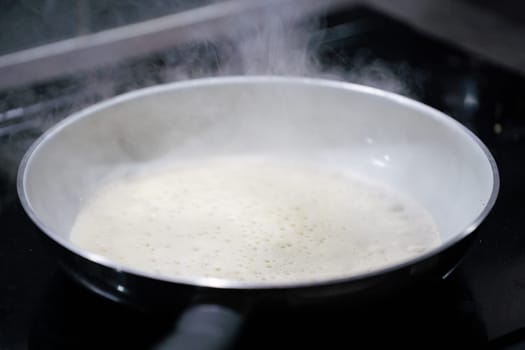 A large frying pan stands on the induction cooker and inside it is a pancake dough that is fried. The water evaporates from the pan.