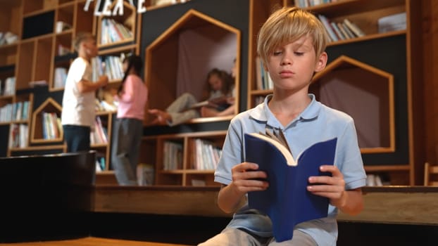 Attractive caucasian boy reading a book while group of smart students sitting at library. Child studying, learning from novel or textbook while children talking, chitchat about education. Erudition.