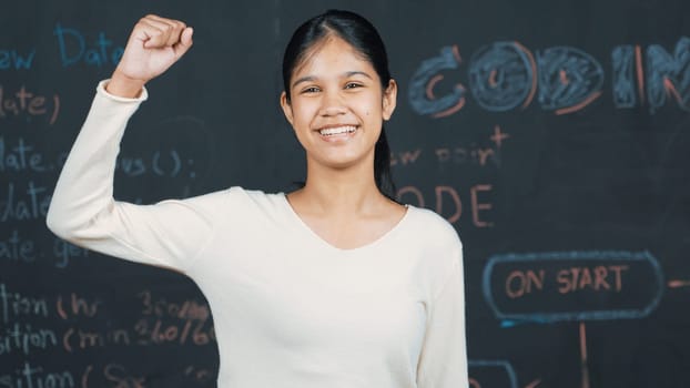 Young student celebrate her successful plan while raise her arm. Happy teenager looking at camera while standing at blackboard with engineering code and prompt written in STEM class. Edification.