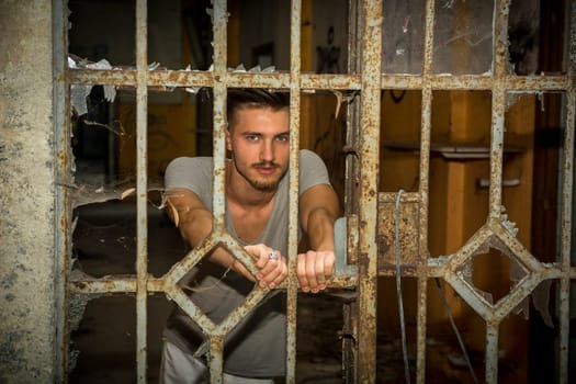 A young blond handsome man standing behind bars in an abandoned house, looking out